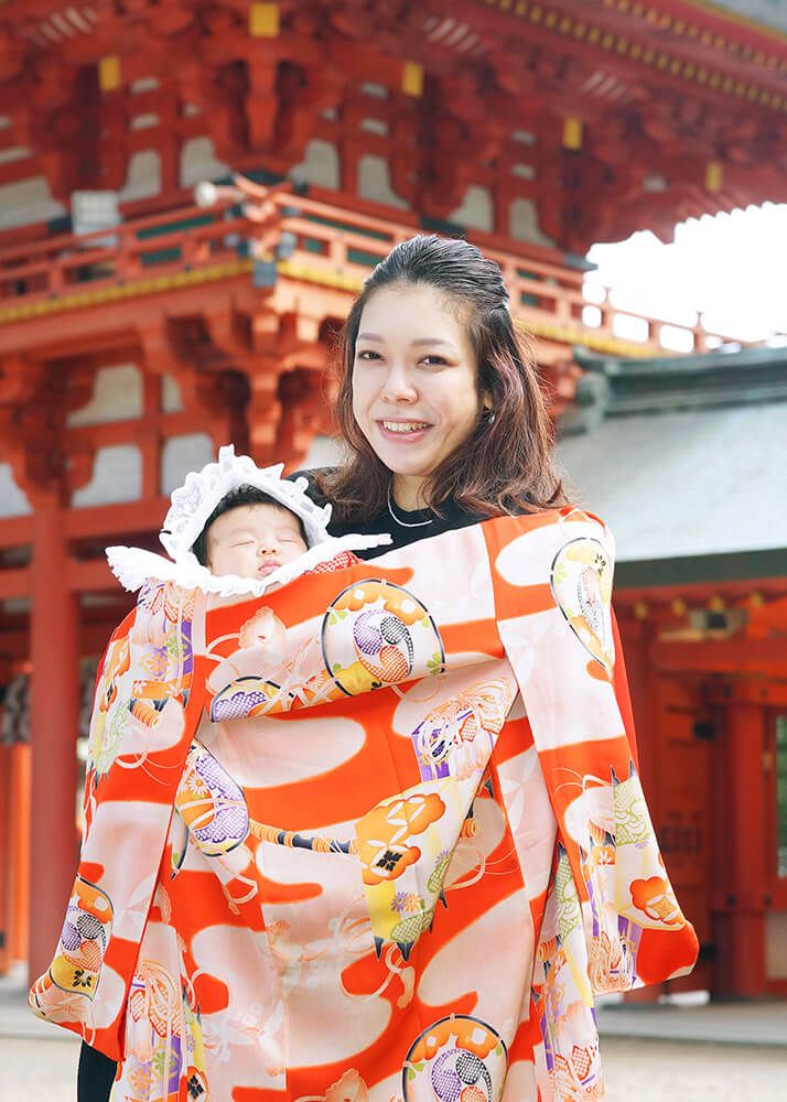大宮氷川神社の楼門前でオレンジ色の祝い着をかけて赤ちゃんを抱っこするママ