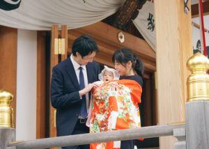 水天宮の本殿前でお宮参りのオレンジ色の祝い着をかけて赤ちゃんを抱っこするママ