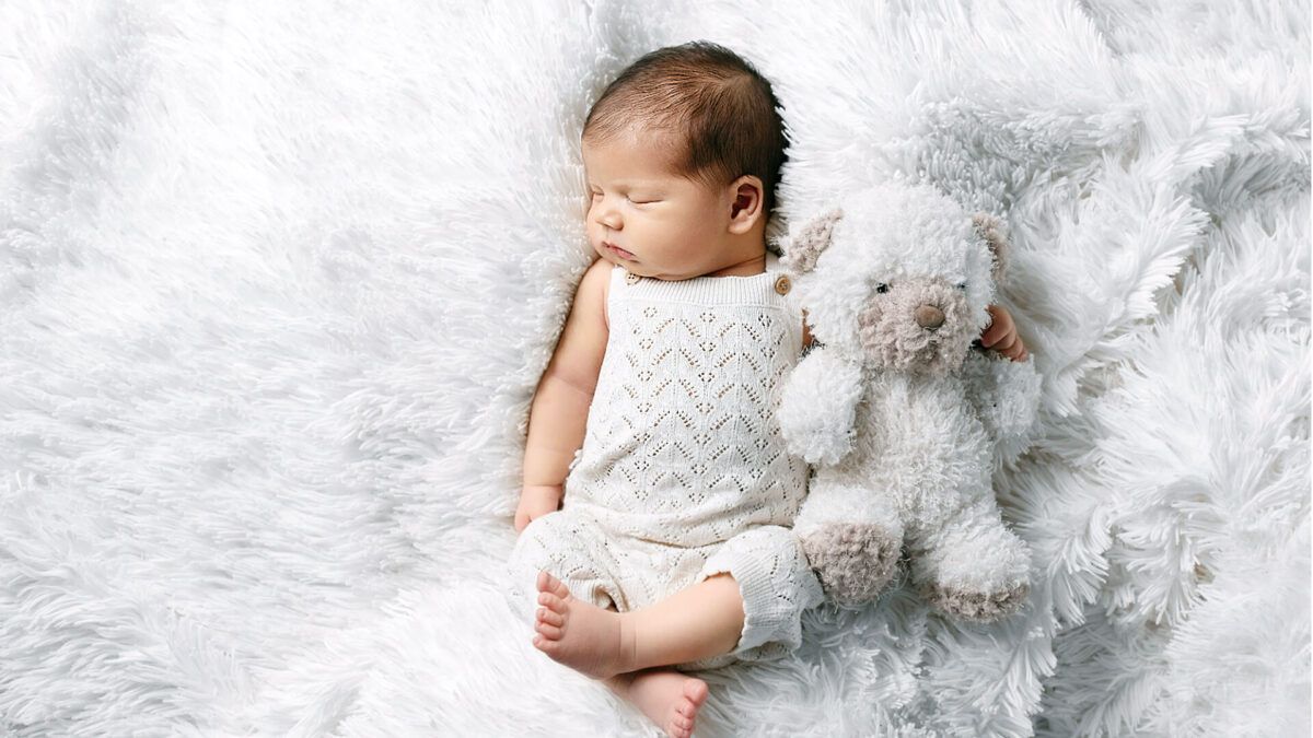 白いファーの上でぬいぐるみを抱えて眠る新生児