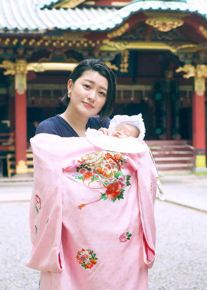 お宮参りでピンク色の祝い着をかけて赤ちゃんを抱っこするママ