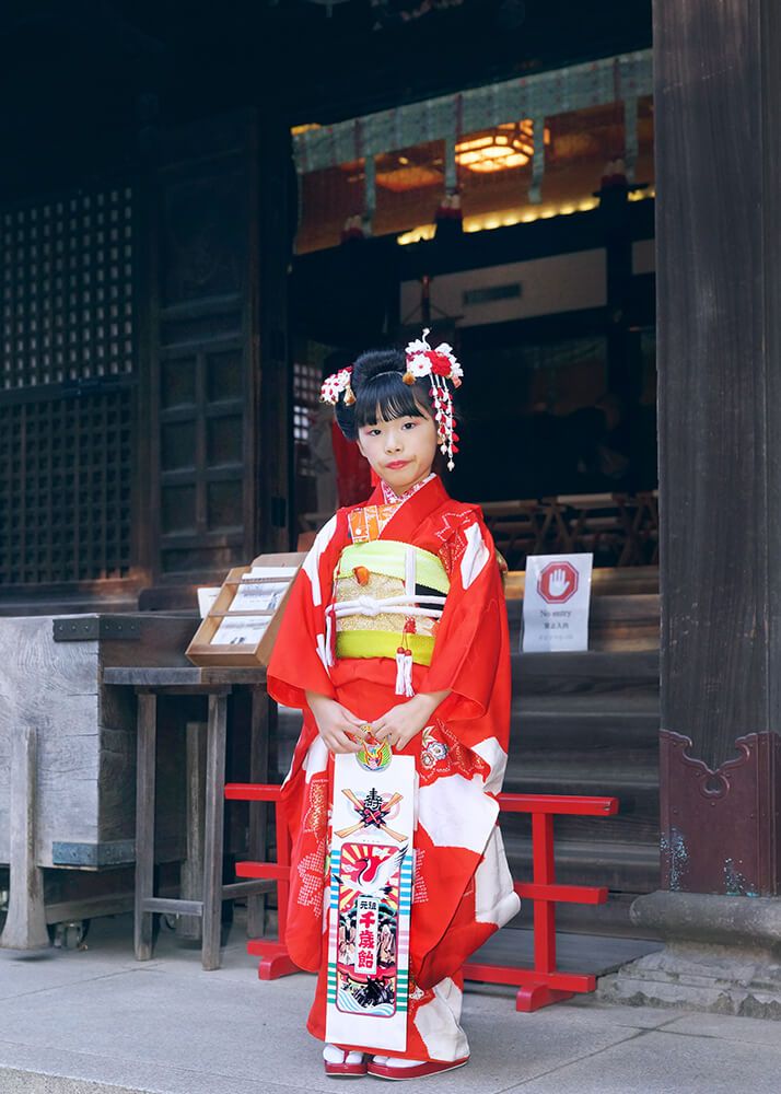 七五三出張撮影で代々木八幡宮の本殿前で千歳飴を持ってポーズをとる7歳女の子の写真