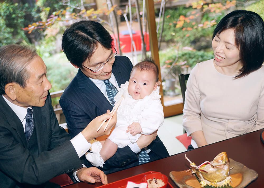 お宮参り・お食い初めロケーションフォト|赤坂日枝神社→芝とうふ屋うかい 17 お食い初めのお膳と祝い鯛を前に並ぶ家族のスナップショットの写真